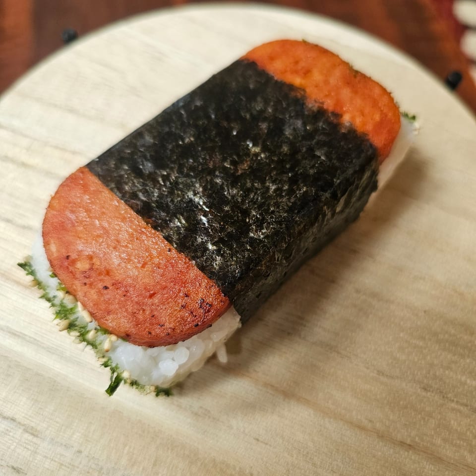 A Kujo Eats Spam Musubi, featuring grilled Spam, rice, and nori, on a wooden board.