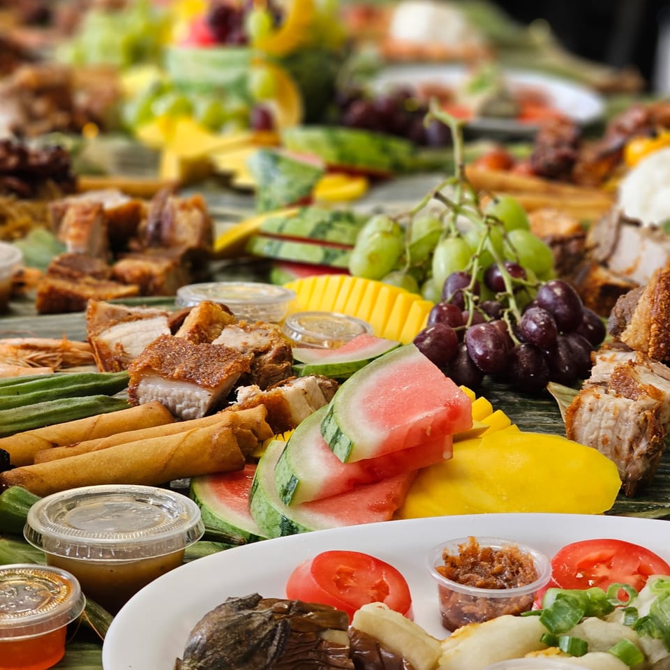 A Filipino boodle fight spread with crispy pork belly, lumpia, watermelon, mango, grapes, and various sauces.