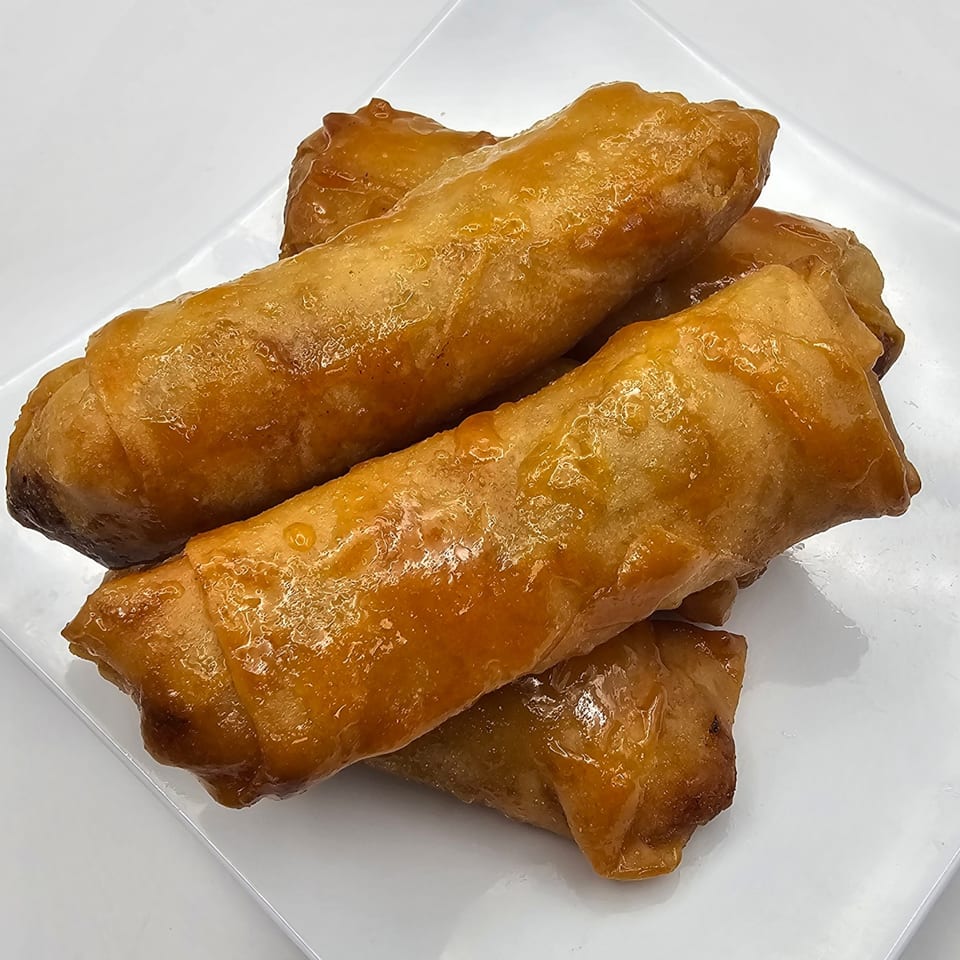 A close-up of several golden-brown, glazed Filipino lumpia on a white plate.