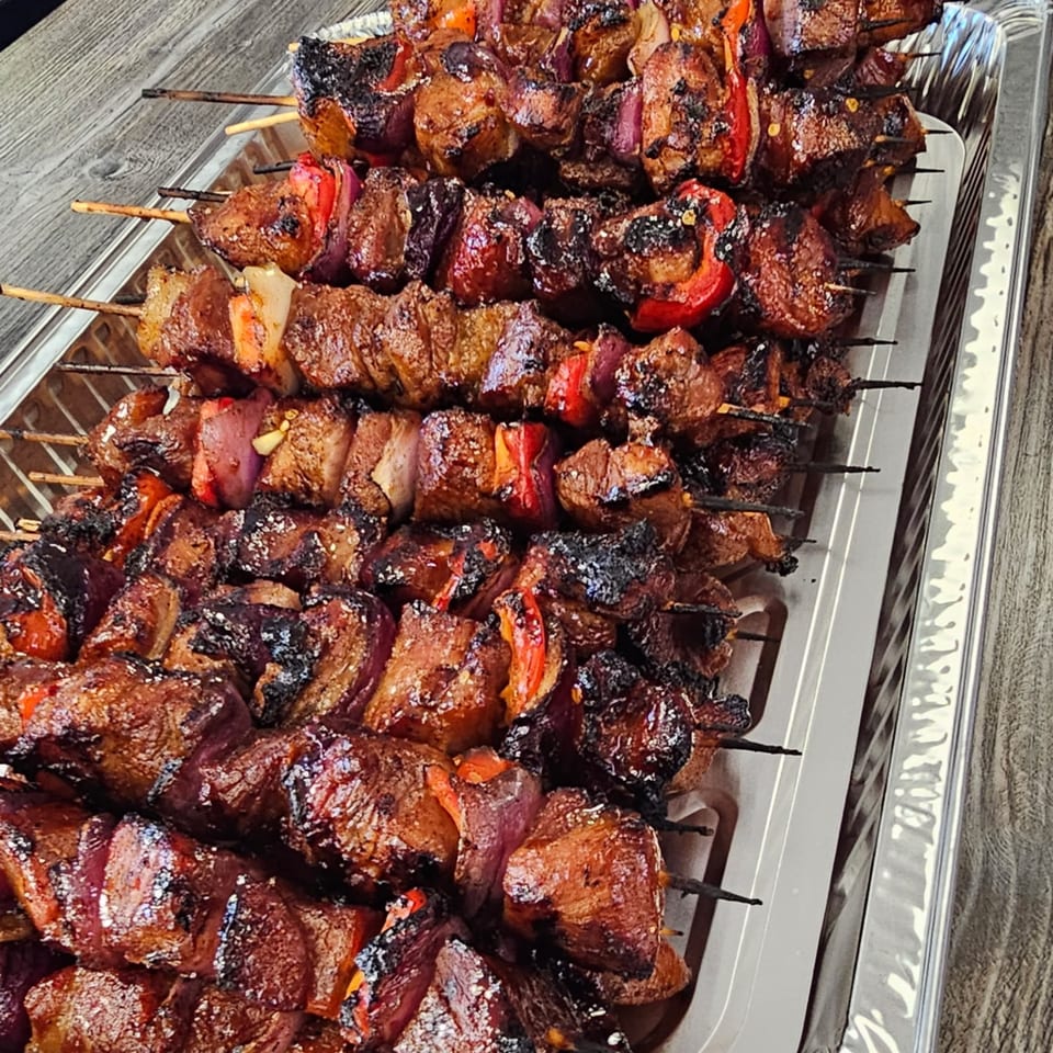 Close-up of Kujo Eats' Filipino BBQ pork skewers with red bell peppers and onions in a foil tray.